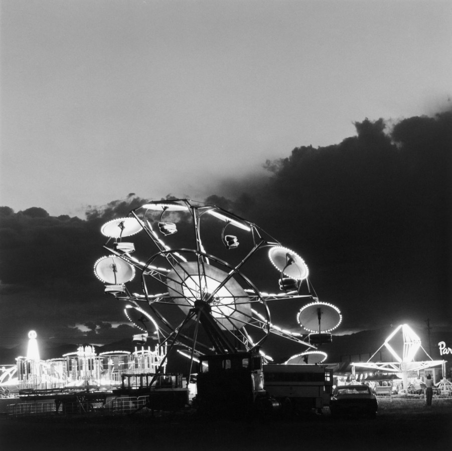 A black and white photograph of an illuminated carnival ride at dusk against a cloudy sky.