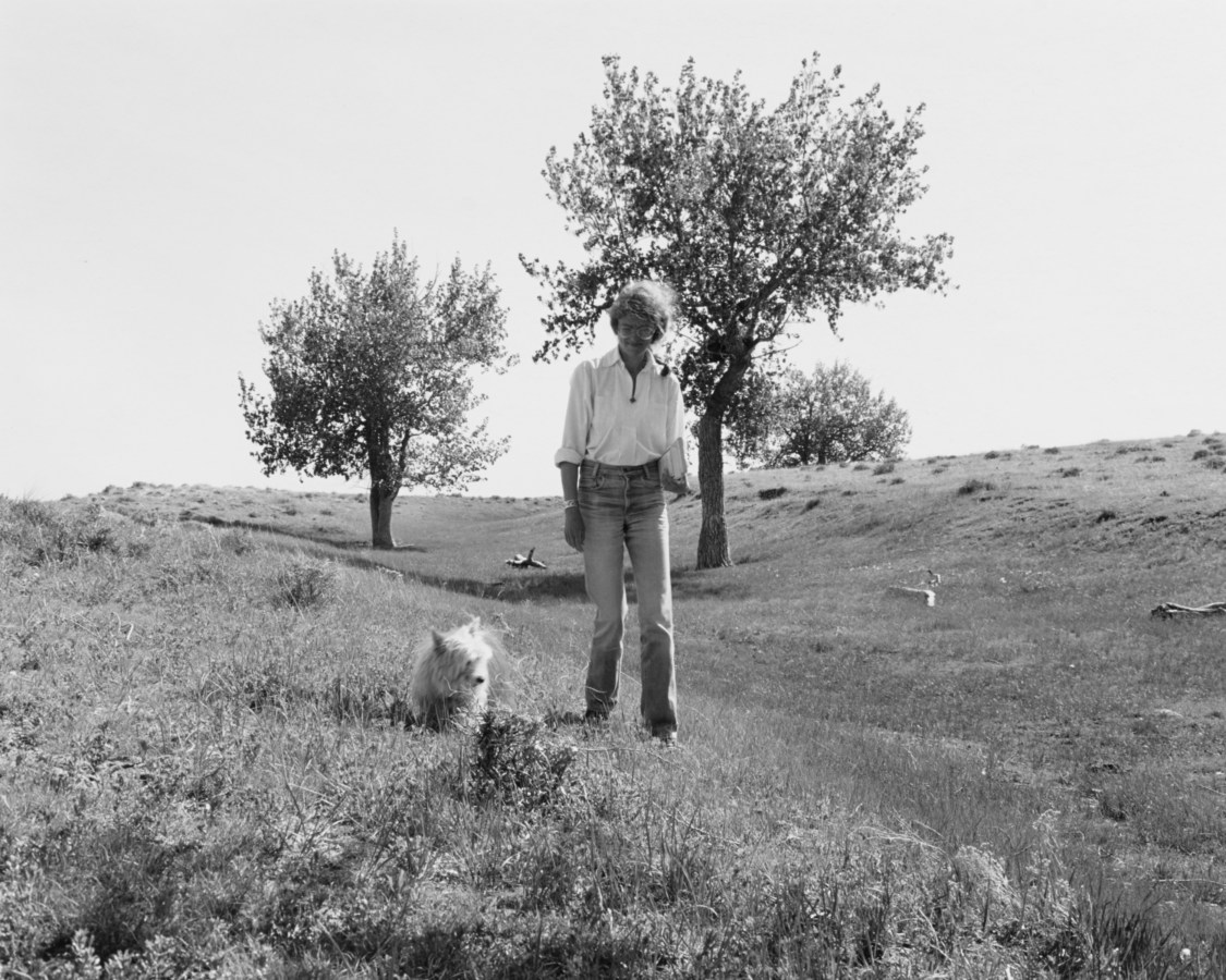 A black and white photograph of a woman and a small white dog trees in the background.