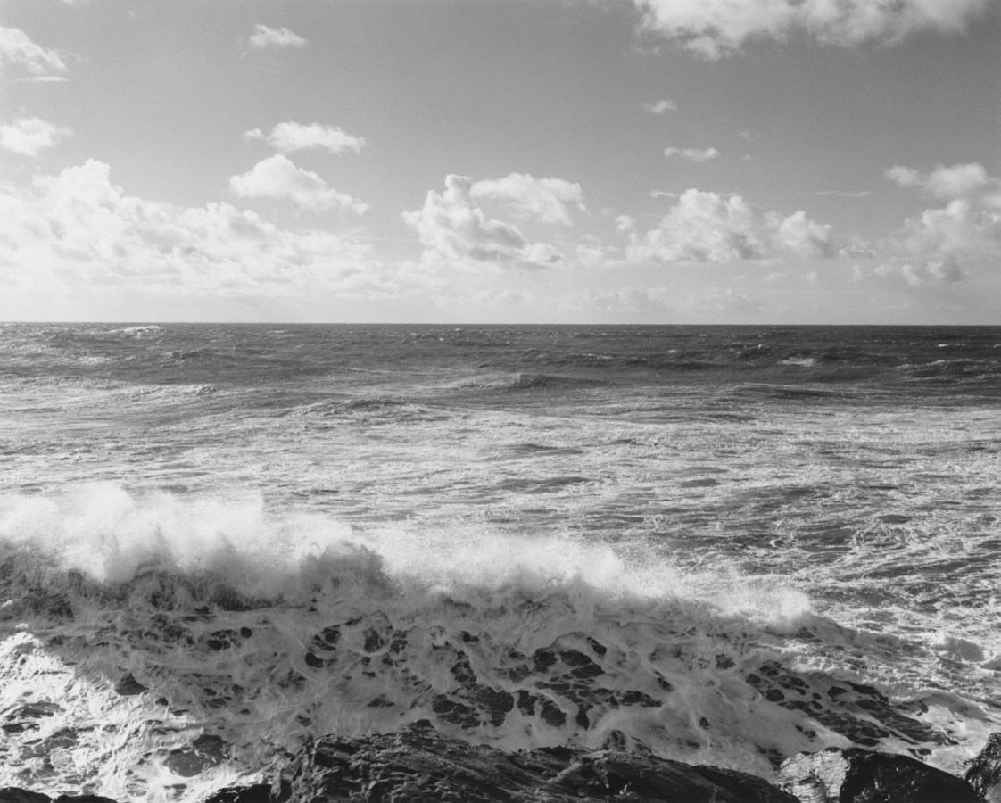 Black-and-white photograph of a wave breaking with scattered clouds in the sky.