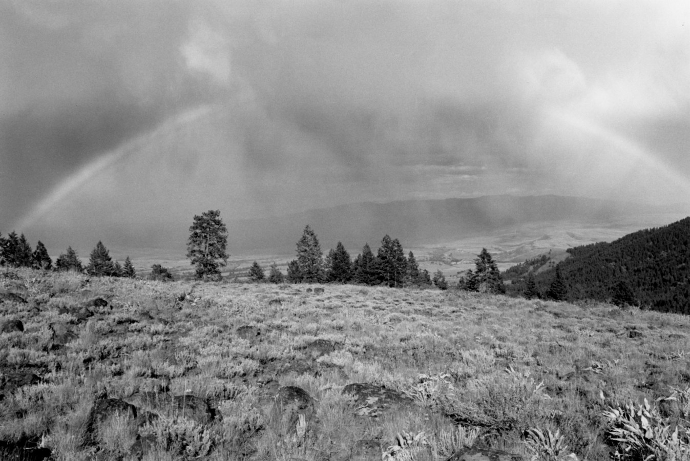 Black-and-white photograph of an open field with trees on the horizon and clouds and a rainbow in the sky