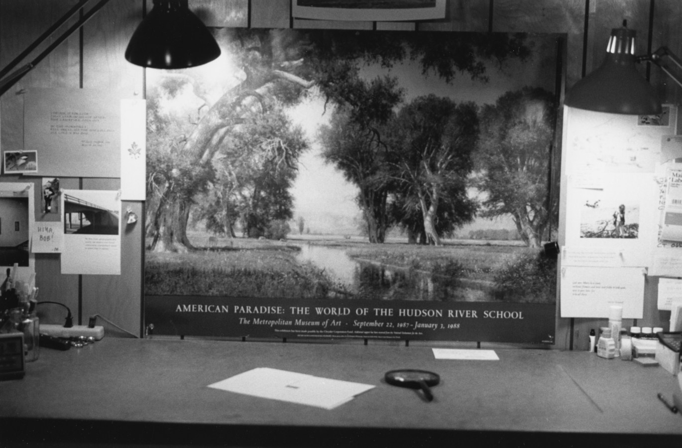 Black-and-white horizontal photograph of a desk with papers and a magnifying glass on the surface and a poster for the "Hudson River School" in the background