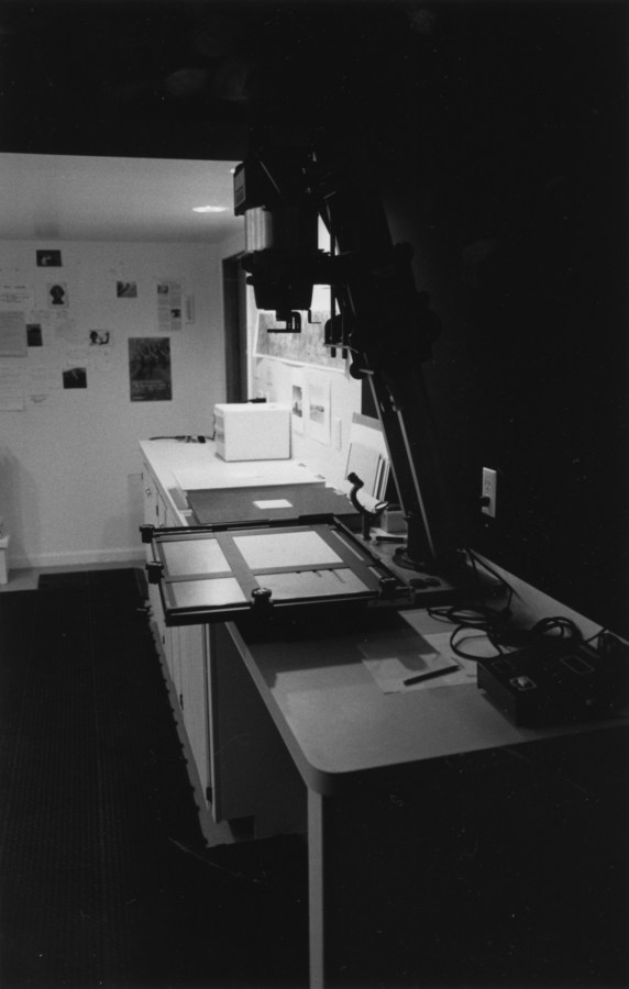 Black-and-white vertical photograph of a photographic dark room showing an enlarger and photographs on the wall