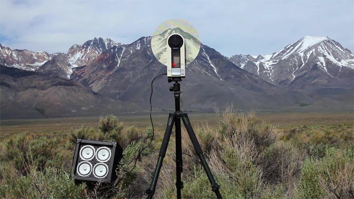 Color film still depicting a tape player on a tripod attached to an amp in middle of grassy field with mountain range