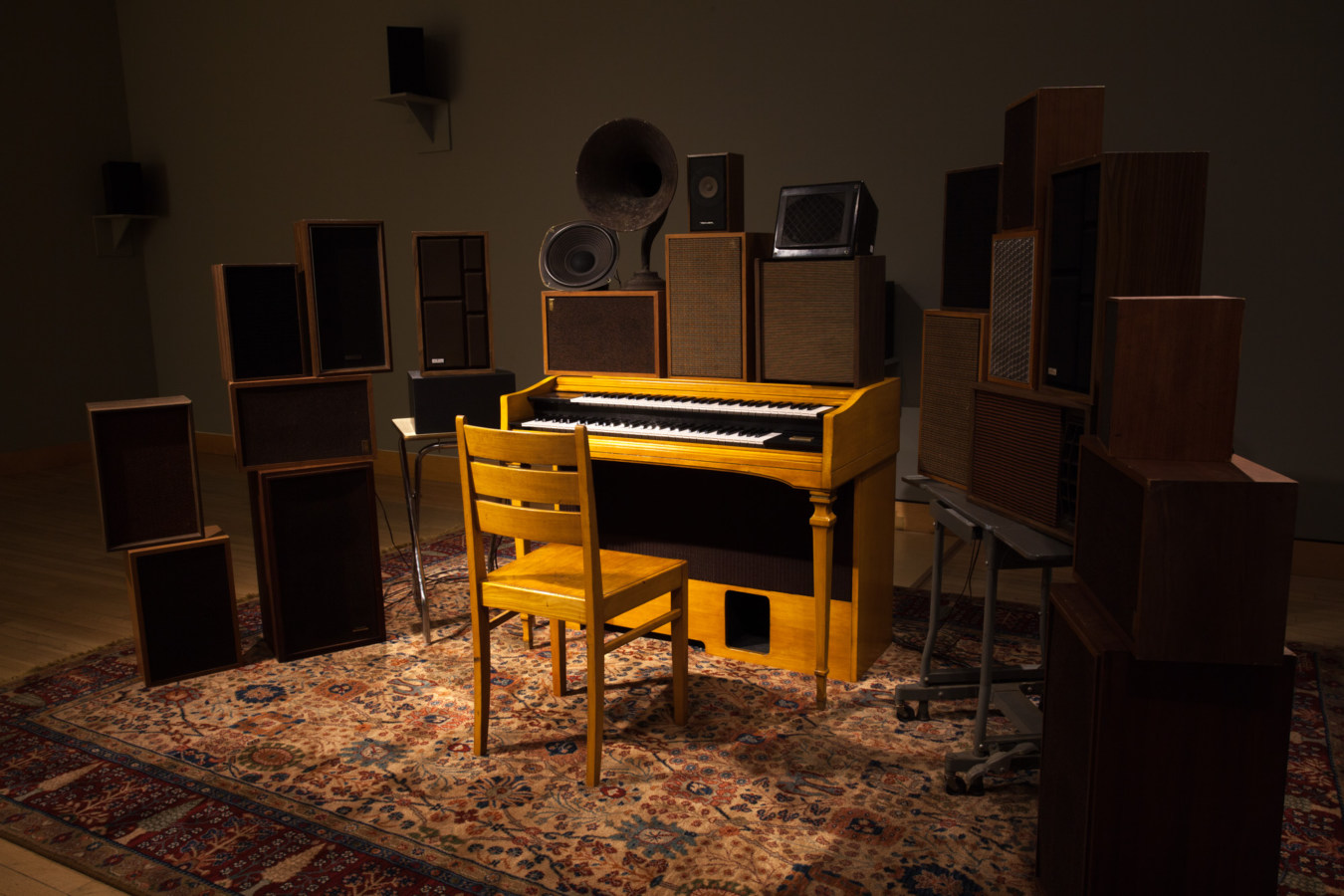 Color image of an organ surrounded by speakers of various sizes and vintages, and an empty chair on an ornate carpet