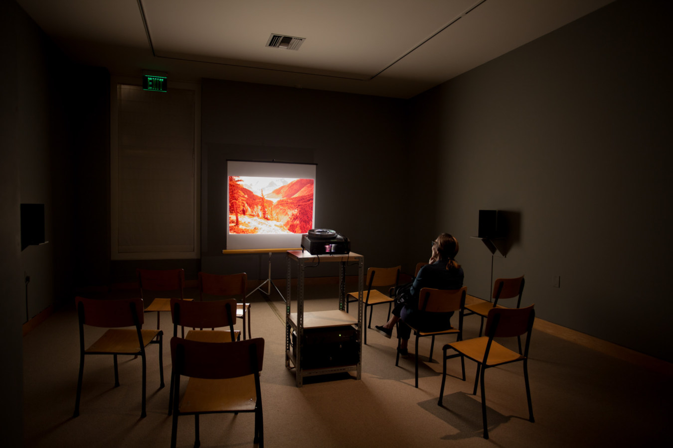 Color image of grey walled gallery with seated person viewing projections and a slide projector exhibiting images of landscapes
