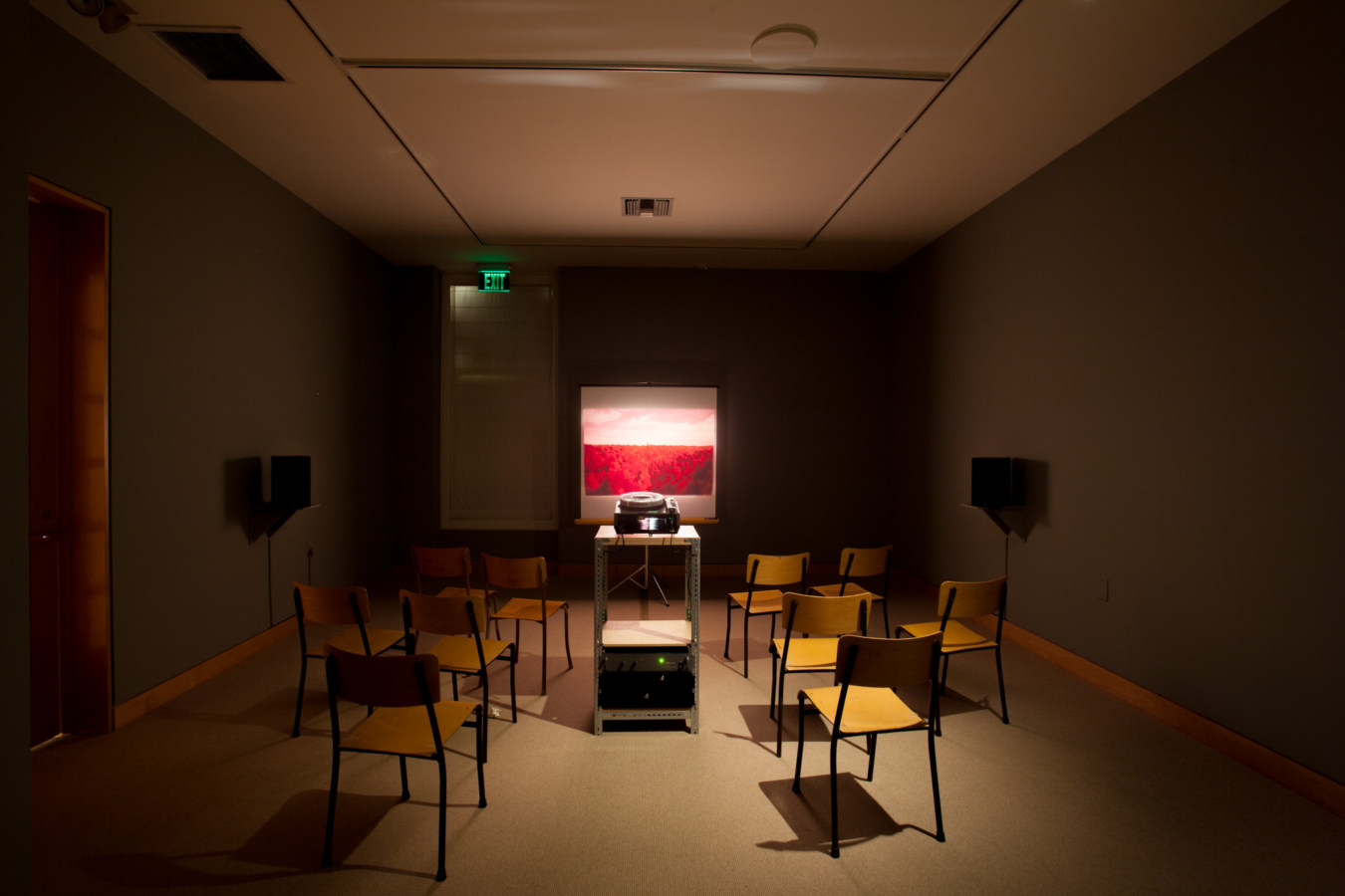 Color image of chairs and slide projector exhibiting slides of landscapes onto white screen within grey gallery walls