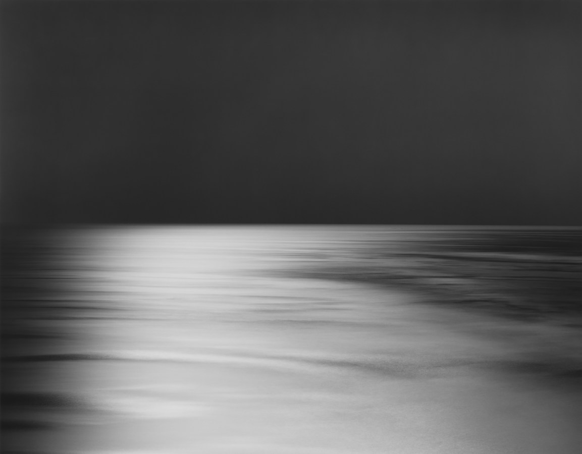 Black-and-white photograph of a seascape, with a black sky and a white light cast across an otherwise black sea
