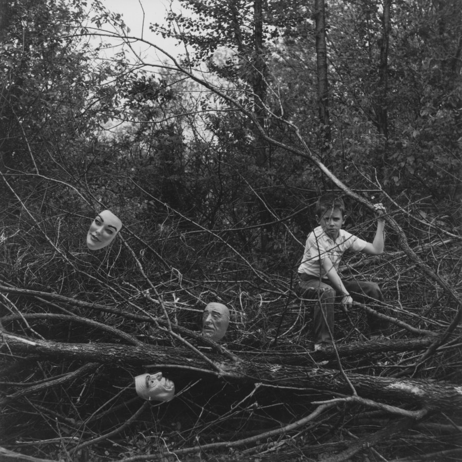 Black-and-white photograph of a boy seated among felled tree branches containing three masks scattered within them