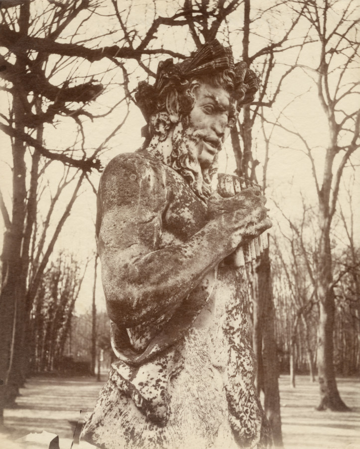 Black and white photograph of a statue depicting a faune holding a pan-flute in a wooded area