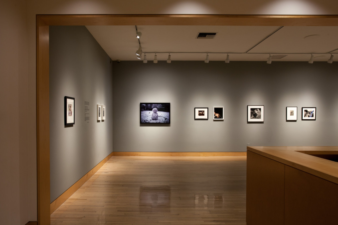 Color image of gallery entryway exhibiting framed color and black and white photographs on grey gallery walls
