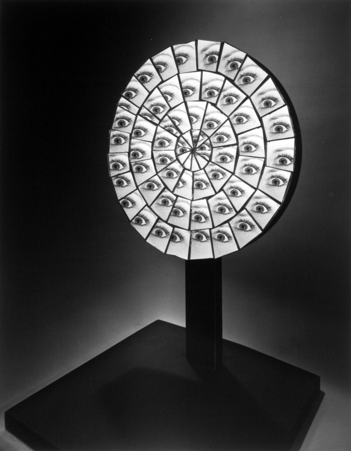 Black and white photograph of circular gridded mirror on black stand with multiple eyes on mirror