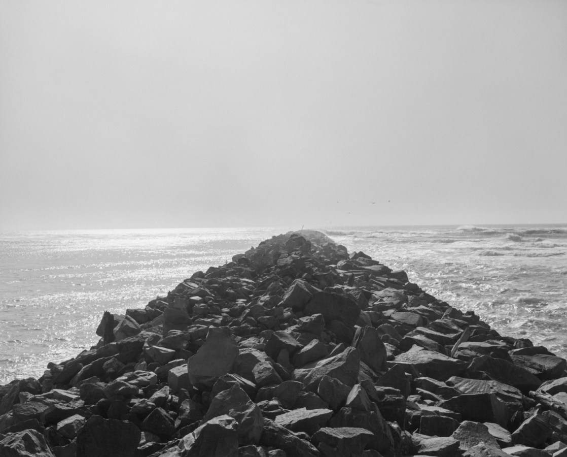 Black-and-white photograph of a rocky jetty leading to the ocean with water on each side.