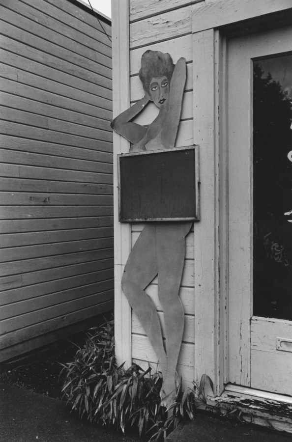 Black and white photograph of building exterior with wooden cutout of women posing seductively with chalkboard covering mid section