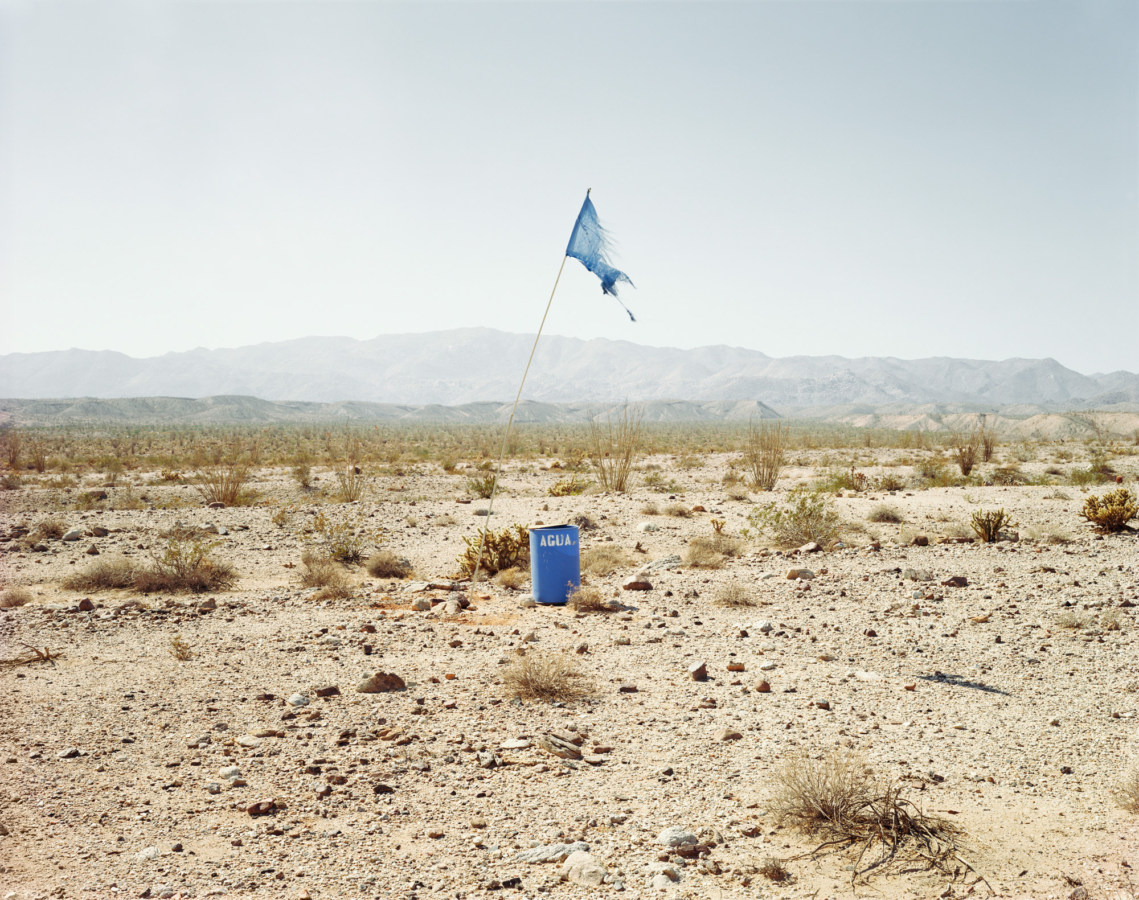 Color photograph of a tattered blue flag standing next to a blue barrel marked AGUA in a desert landscape