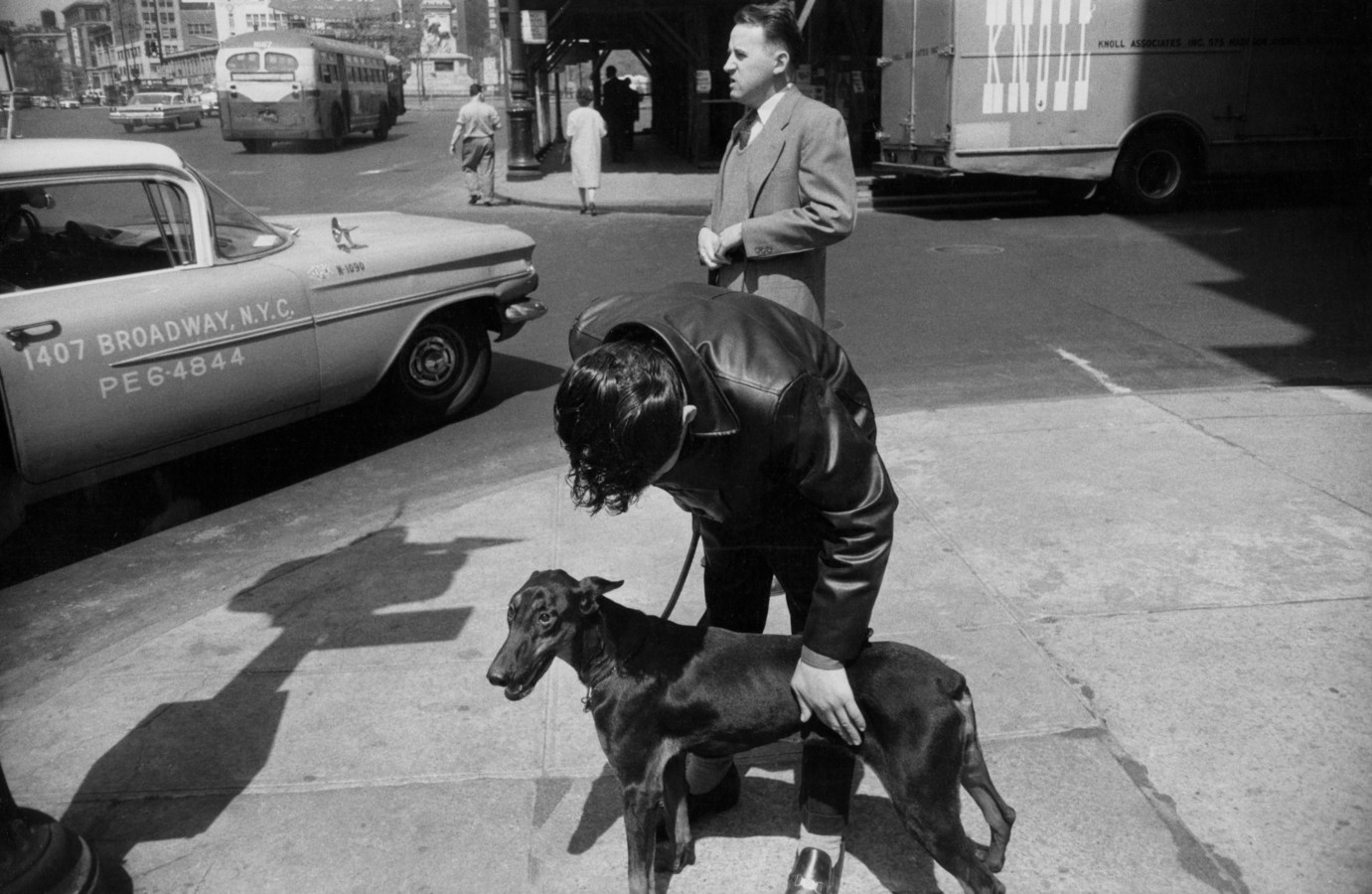 Black and white photograph of two men on the street, one leaning over a medium-sized black dog