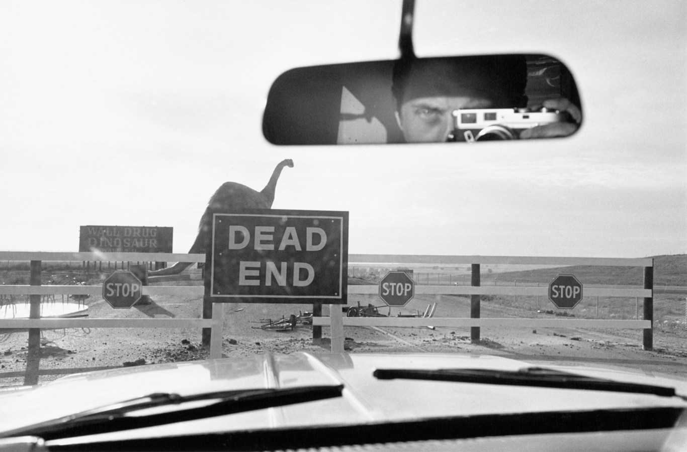 Black and white photograph of sign reading "Dead End" taken from interior of car with large dinosaur in empty desert in background
