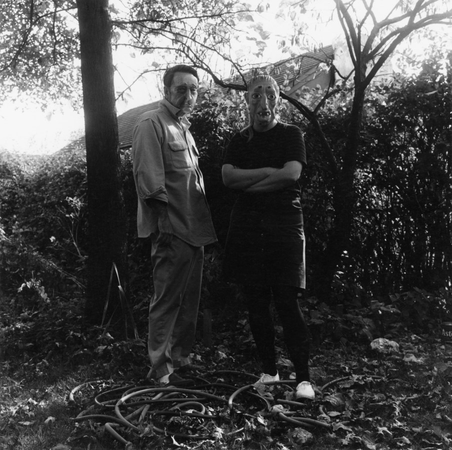 Black-and-white photograph of two people in rubber masks standing against a hedgerow