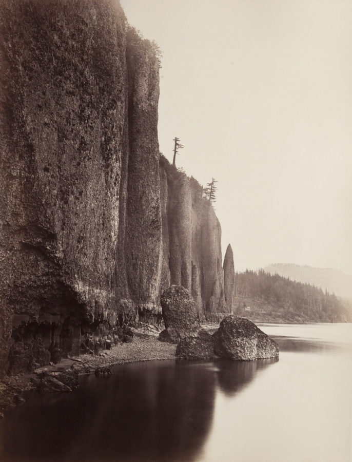 Color photograph of a cliffside and water with trees on horizon