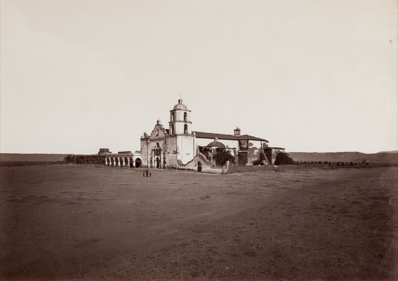 Black and white photograph of exterior of mission in middle of desert valley