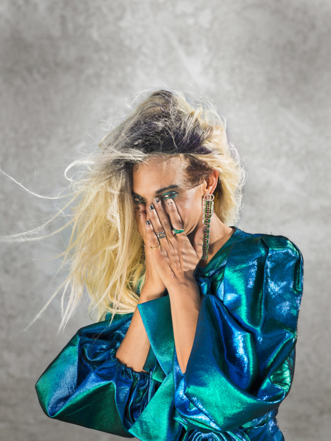 Color photographic portrait of a woman in an iridescent blue top covering her face with her hands