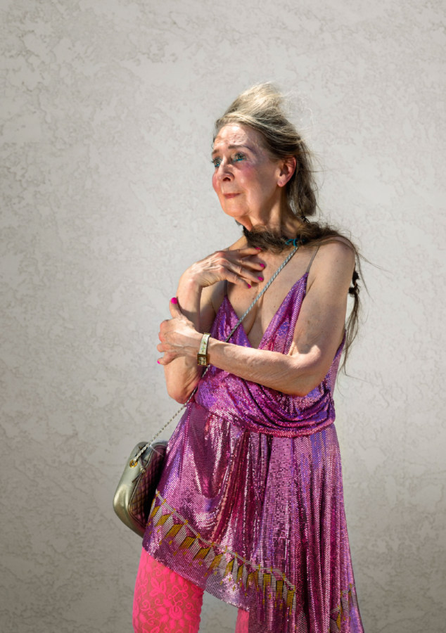 Color photographic portrait of an older woman in a pink sparkly top with her hand on her chest gazing off camera