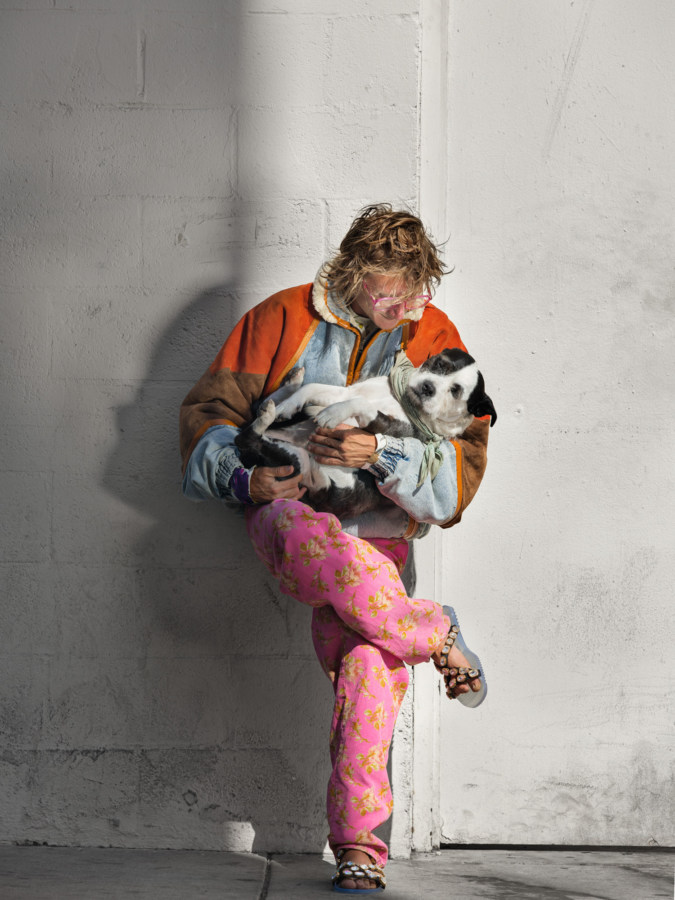 Color photograph of a man in a colorful mismatched outfit holding a black and white dog while leaning on a white wall