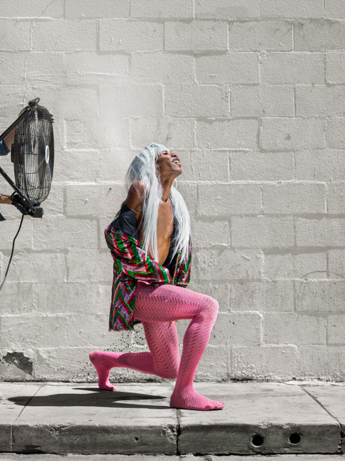 Color photograph of a woman in pink tights lunging in front of a fan and a cinderblock wall