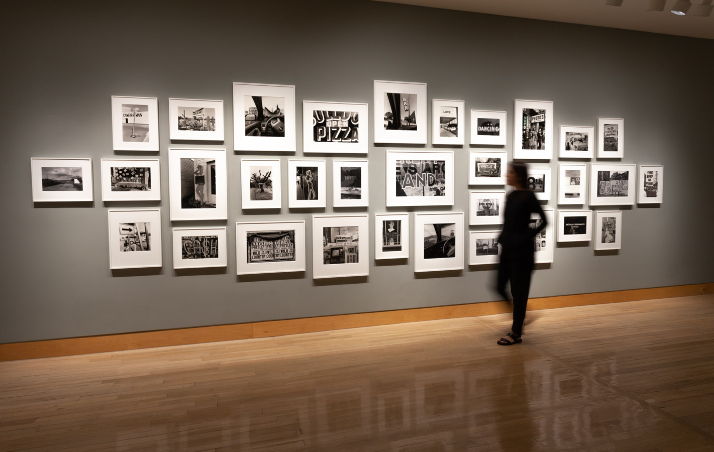 Color image of black and white photographs on grey gallery wall with person viewing the work