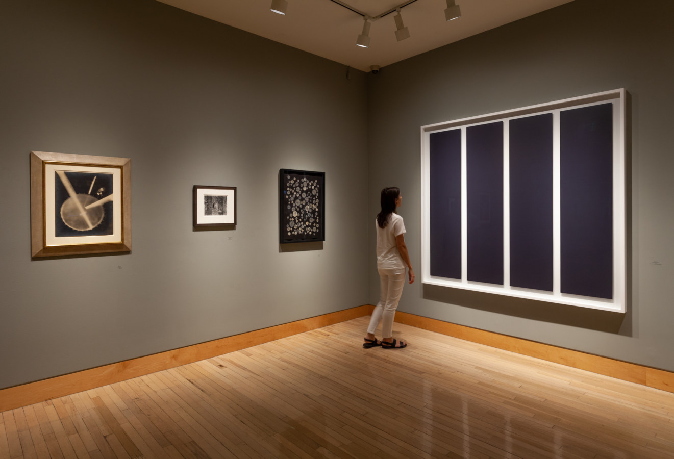 Color image of framed photographs in both color and black and white on grey gallery wall with person viewing work