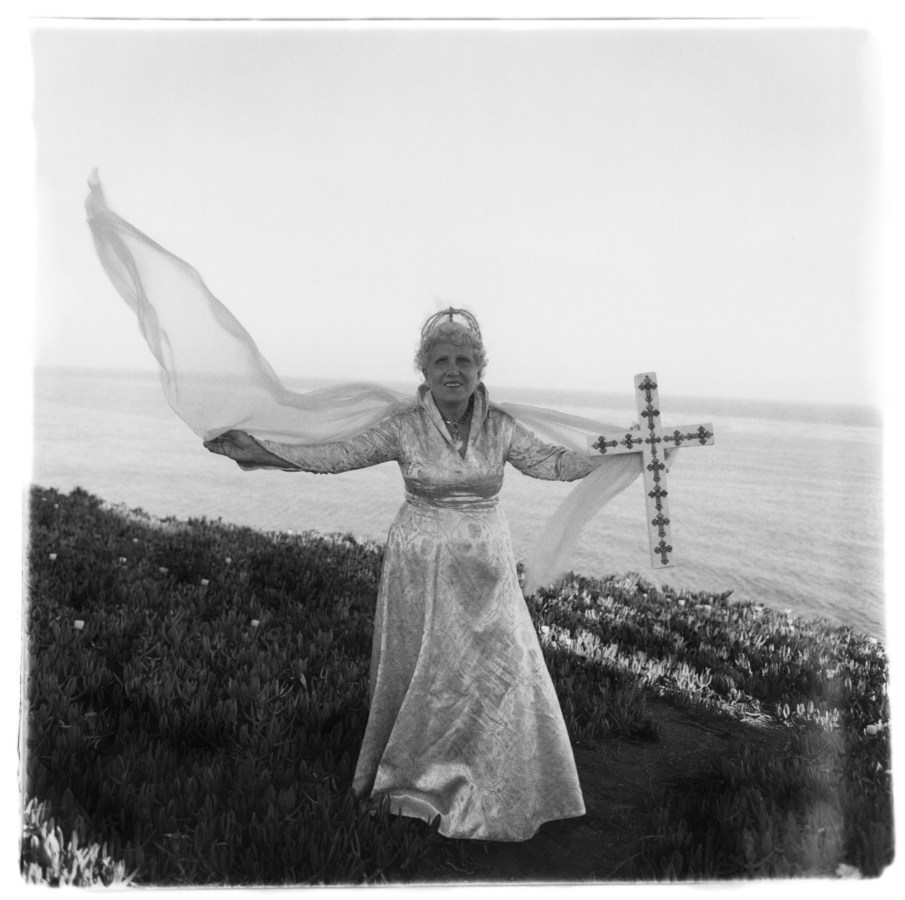 Black-and-white photograph of a woman in a white dress holding a cross, her veil blows in the wind