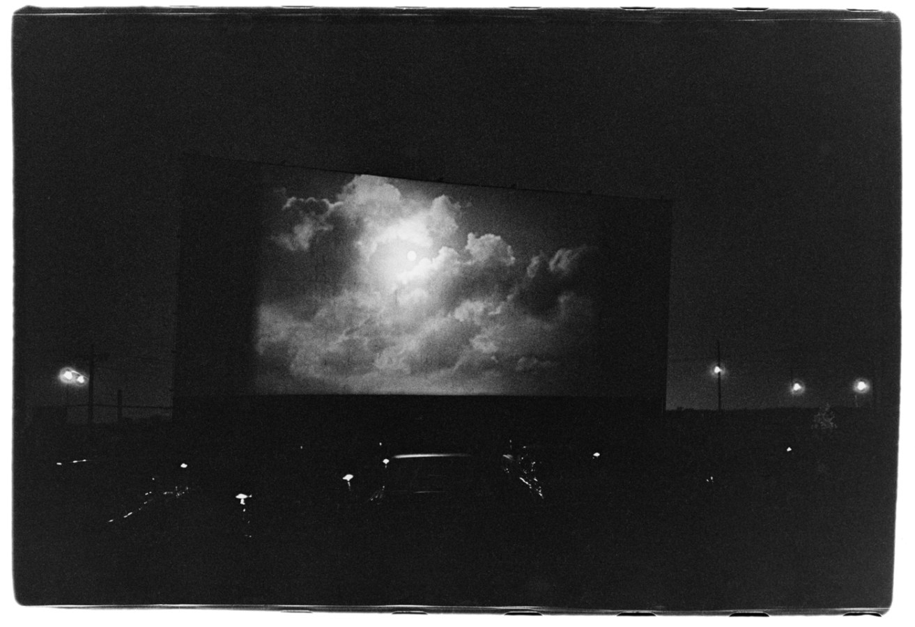 Black-and-white photograph of a drive-in theater at nighttime with clouds on screen