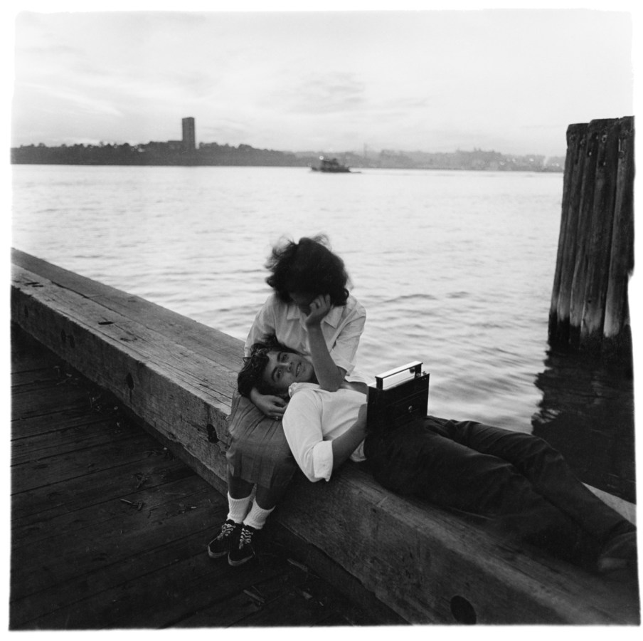 Black-and-white photograph of a woman sitting with man laying down resting his head on her lap with water in the background