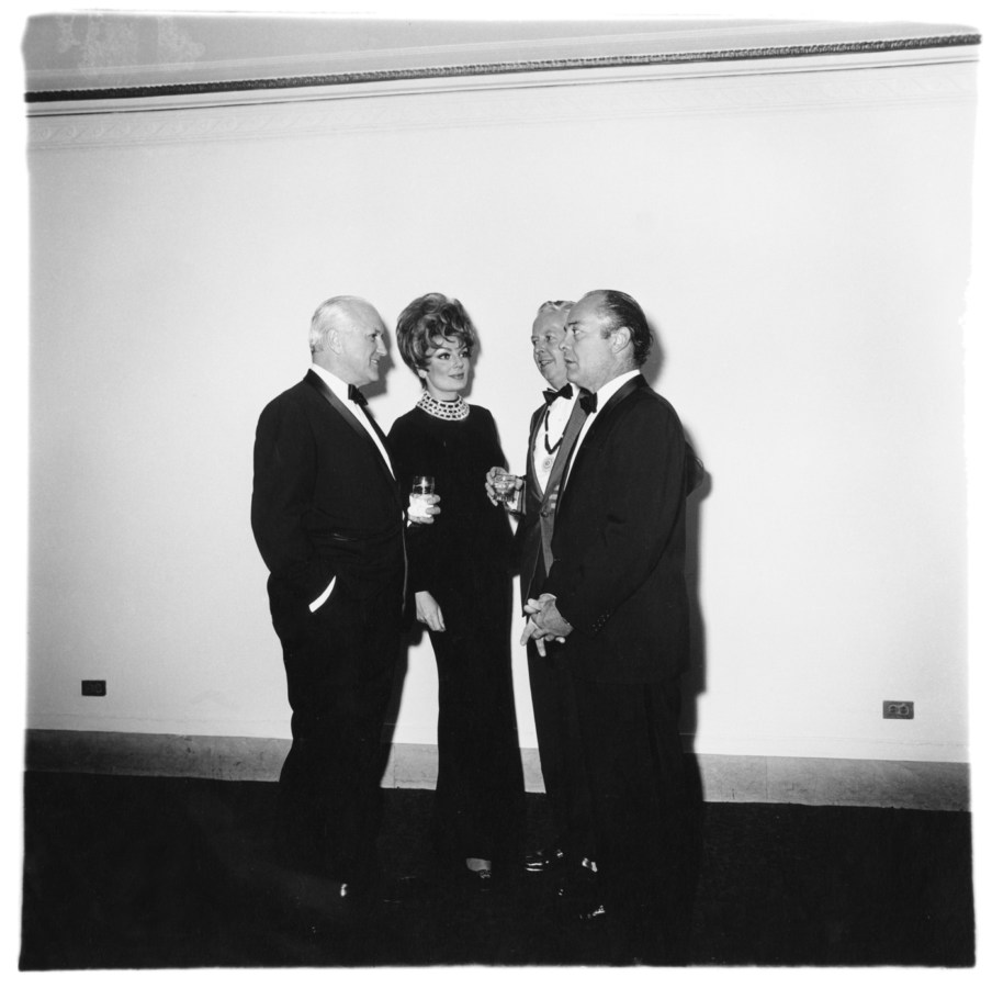 Black-and-white photograph of four figures wearing formal attire gathered in front of a white wall