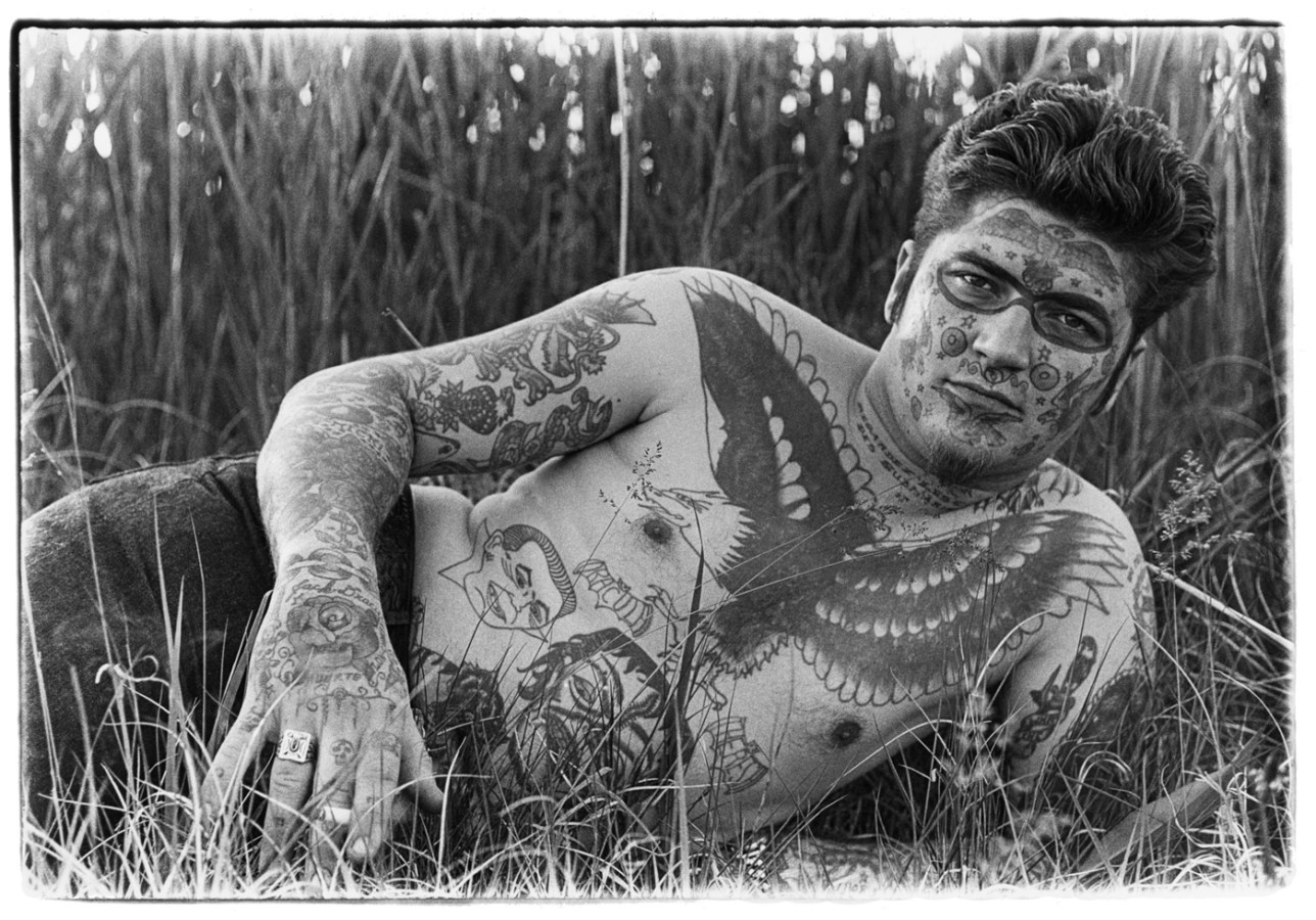 Black-and-white photograph of a shirtless man with tattoos laying in the grass