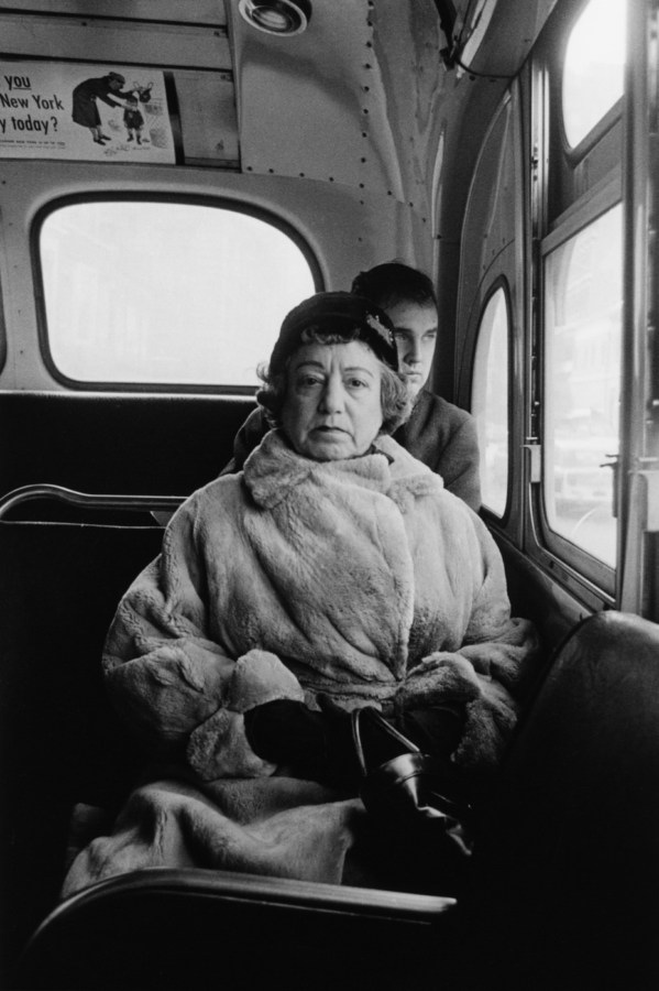 Black-and-white photograph of a woman wearing a fur coat and hat sitting in the back of a bus