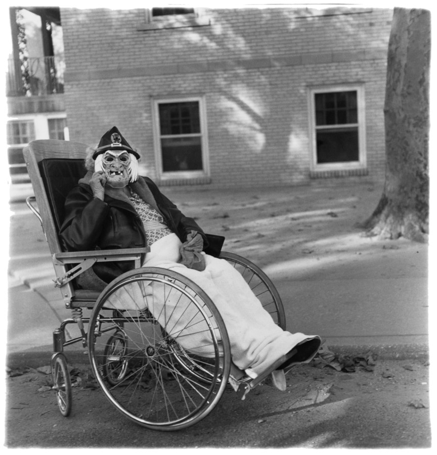 Black-and-white photograph of a masked figure in a wheelchair with a building and tree in the background