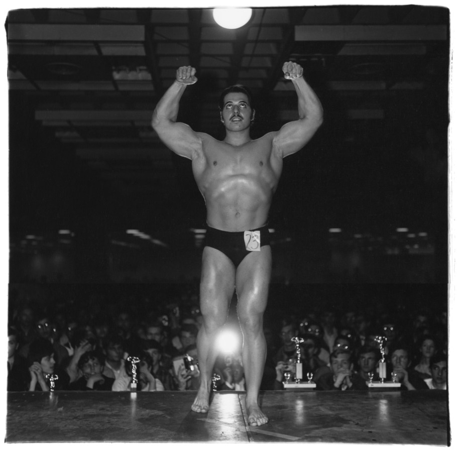 Black-and-white photograph of a body builder flexing on stage with his back to the crowd