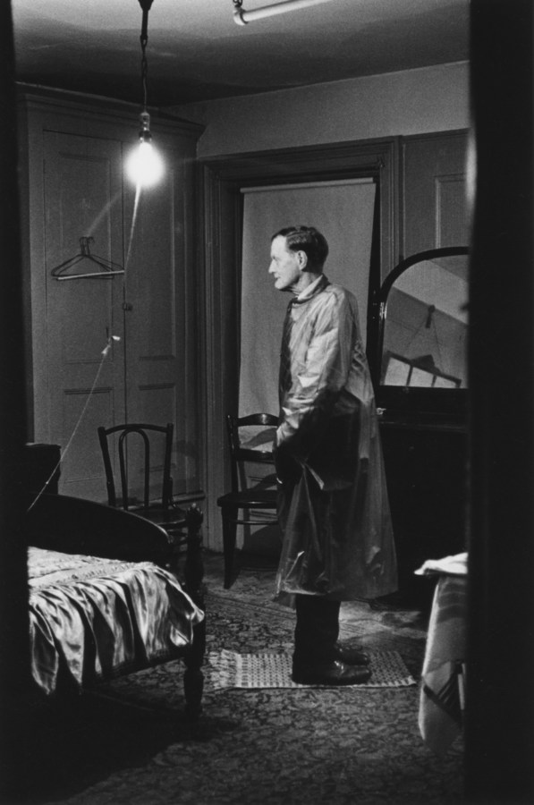 Black-and-white photograph of a man with his head facing the opposite way of his body in a room lit by a single lightbulb