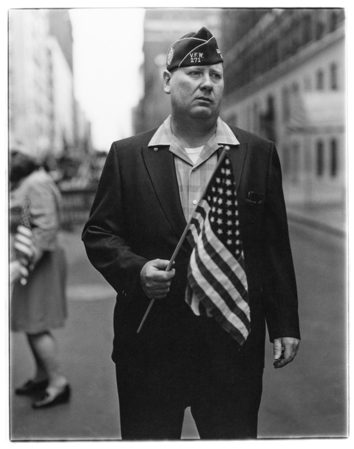Black-and-white photograph of a man standing in a street holding an American flag