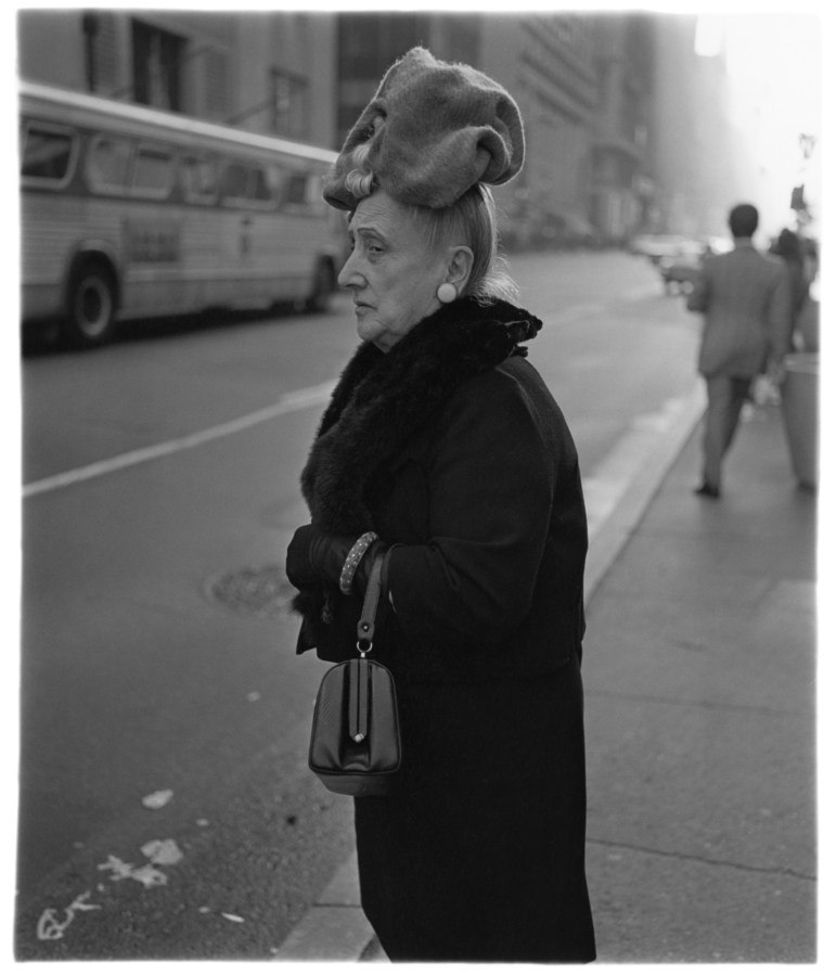 Black-and-white photograph of a woman standing on the edge of the sidewalk with a bus in the background