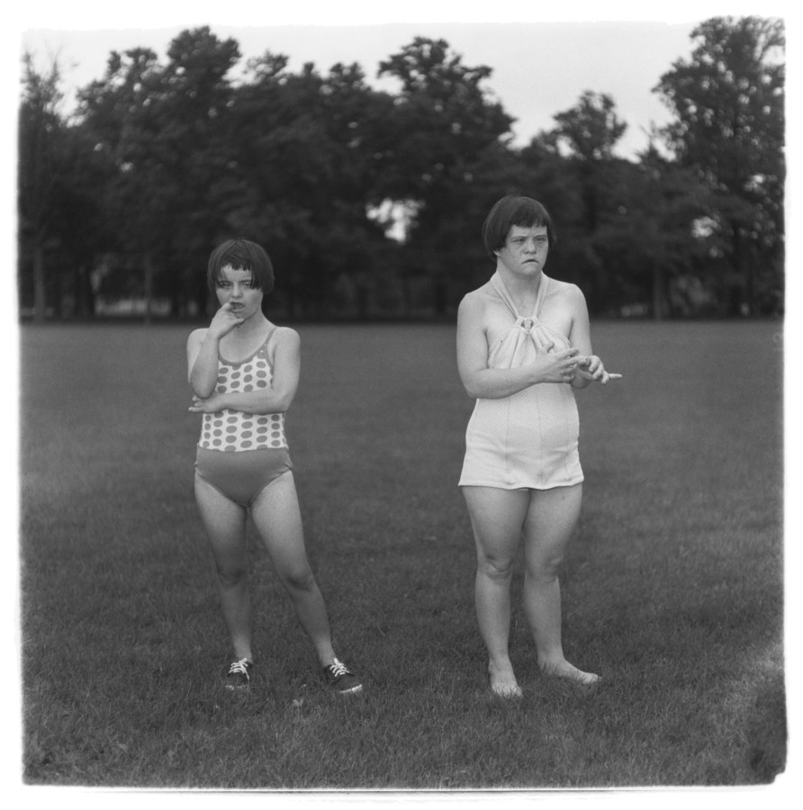 Black-and-white photograph of two figures wearing bathing suits standing in a field with a line of trees in the background
