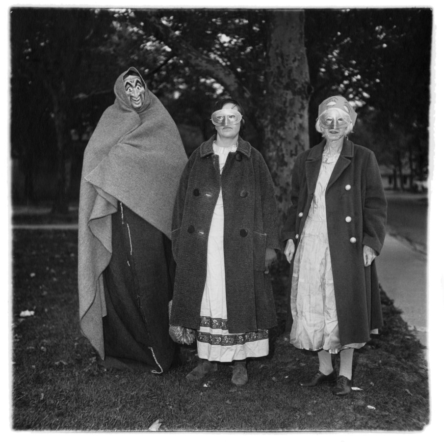 Black-and-white photograph of three masked figures in coats with trees in the background