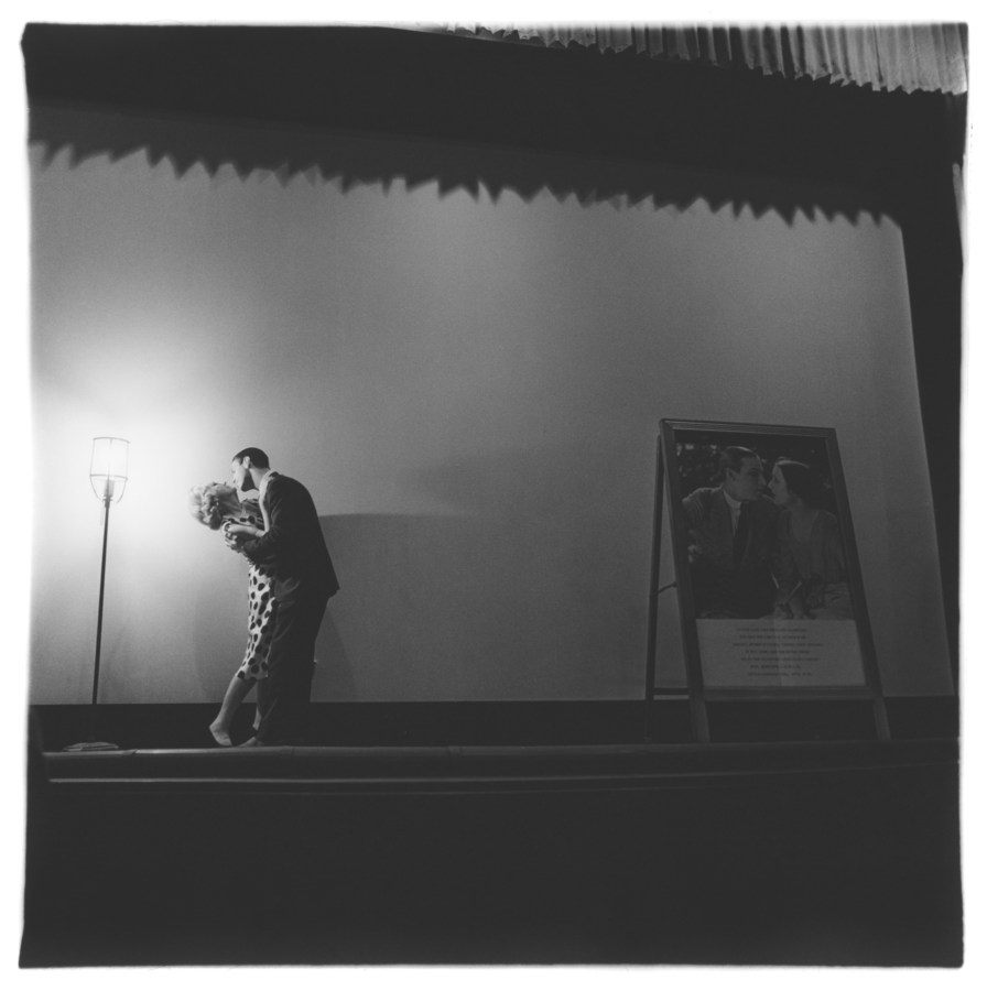 Black-and-white square photograph of two figures in embrace on an empty stage, the their left is a single floor lamp illuminating the scene to their right is a poster depicting a couple kissing