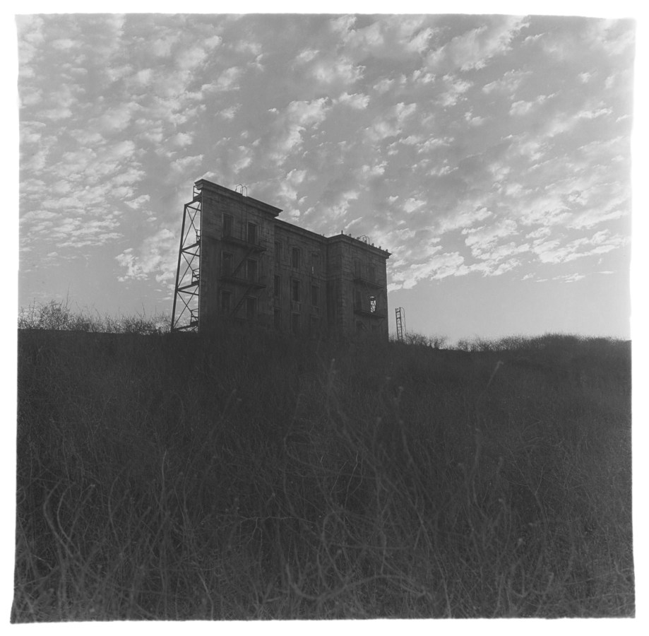 Black-and-white photograph of a fake building facade with grass in the foreground and a scattered clouds in the sky