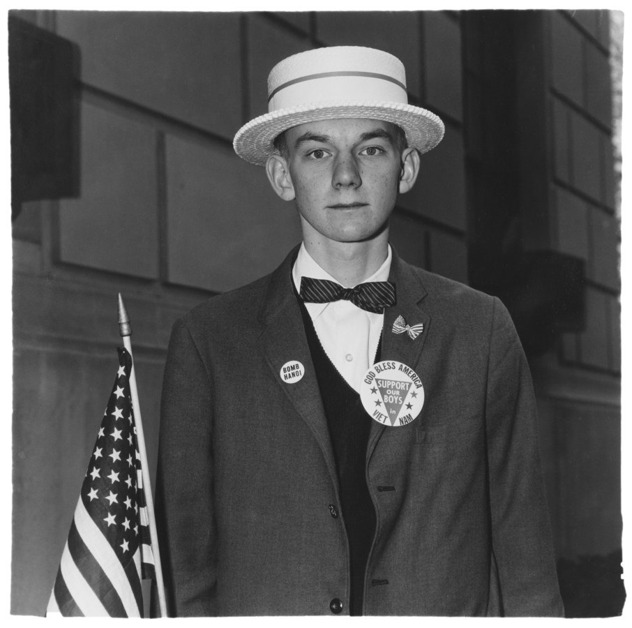 Black-and-white photograph of a man with a hat, bowtie, jacket with political lapel pins holding an American flag