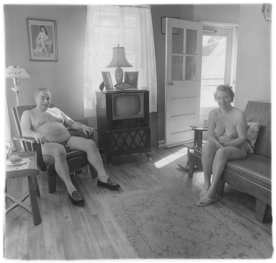 Black-and-white photograph of a nude man and woman sitting in a living room with a television at the center