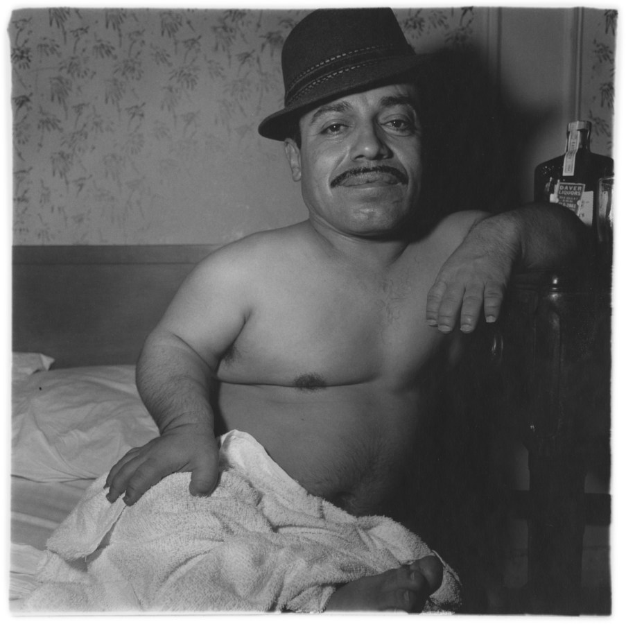 Black-and-white photograph of shirtless man wearing a hat sitting in bed with a towel on his lap