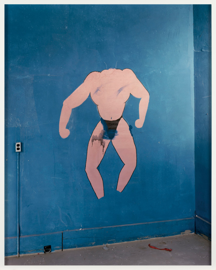 A framed photograph of a blue wall, painted with a picture of a headless muscle man