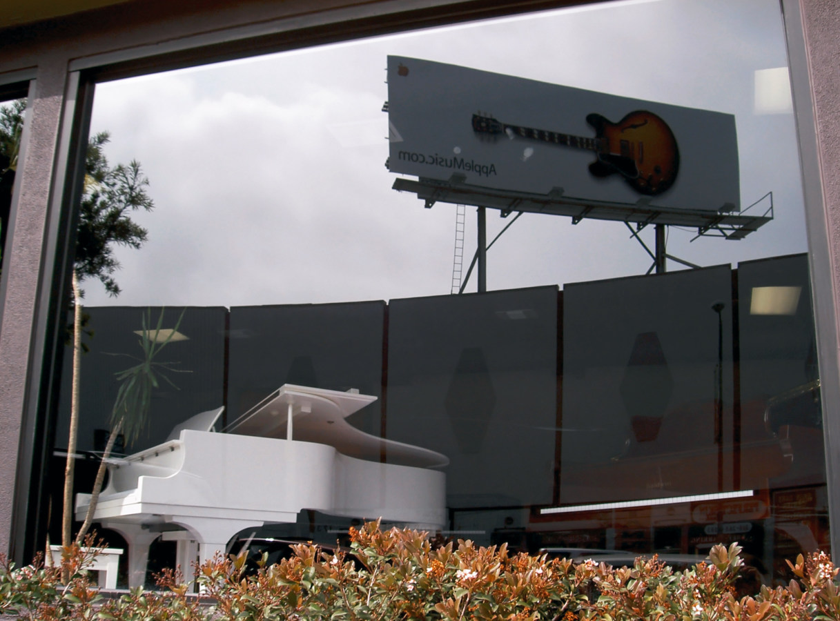 A photograph of a white grand piano seen through a window, upon which is reflected a billboard of a guitar.