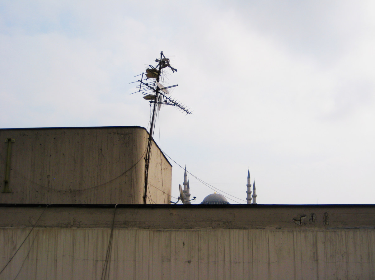 A color photograph of the top of a building, with speakers and satellite dishes on the roof. A mosque peeks out from the other side.